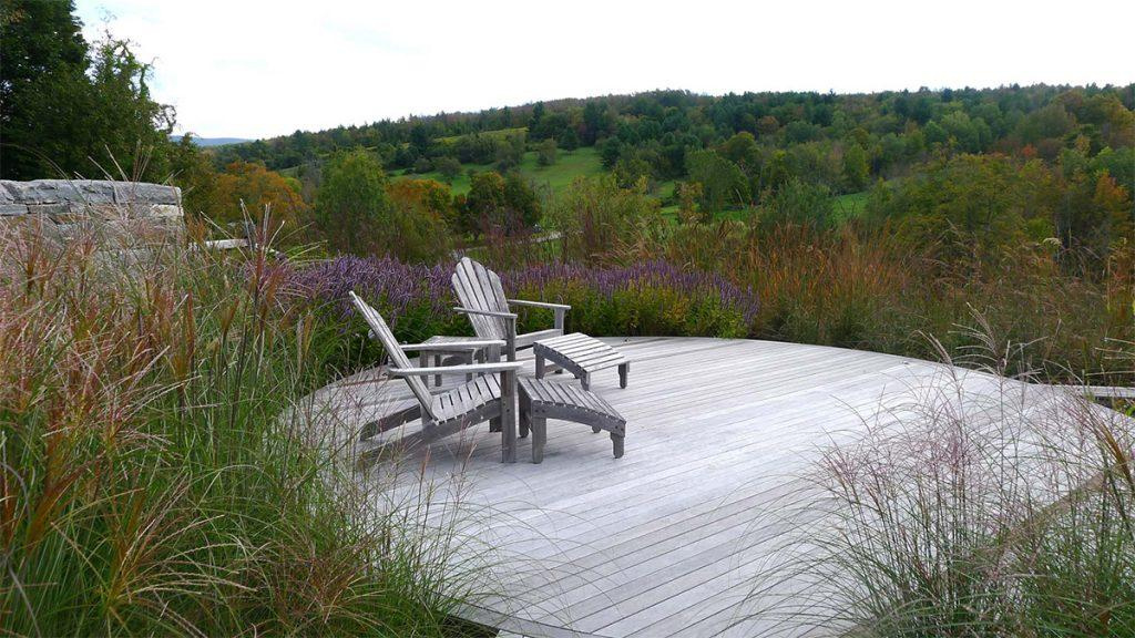 A couple of wooden lounge chairs on a wooden patio overlooking green plants and trees.